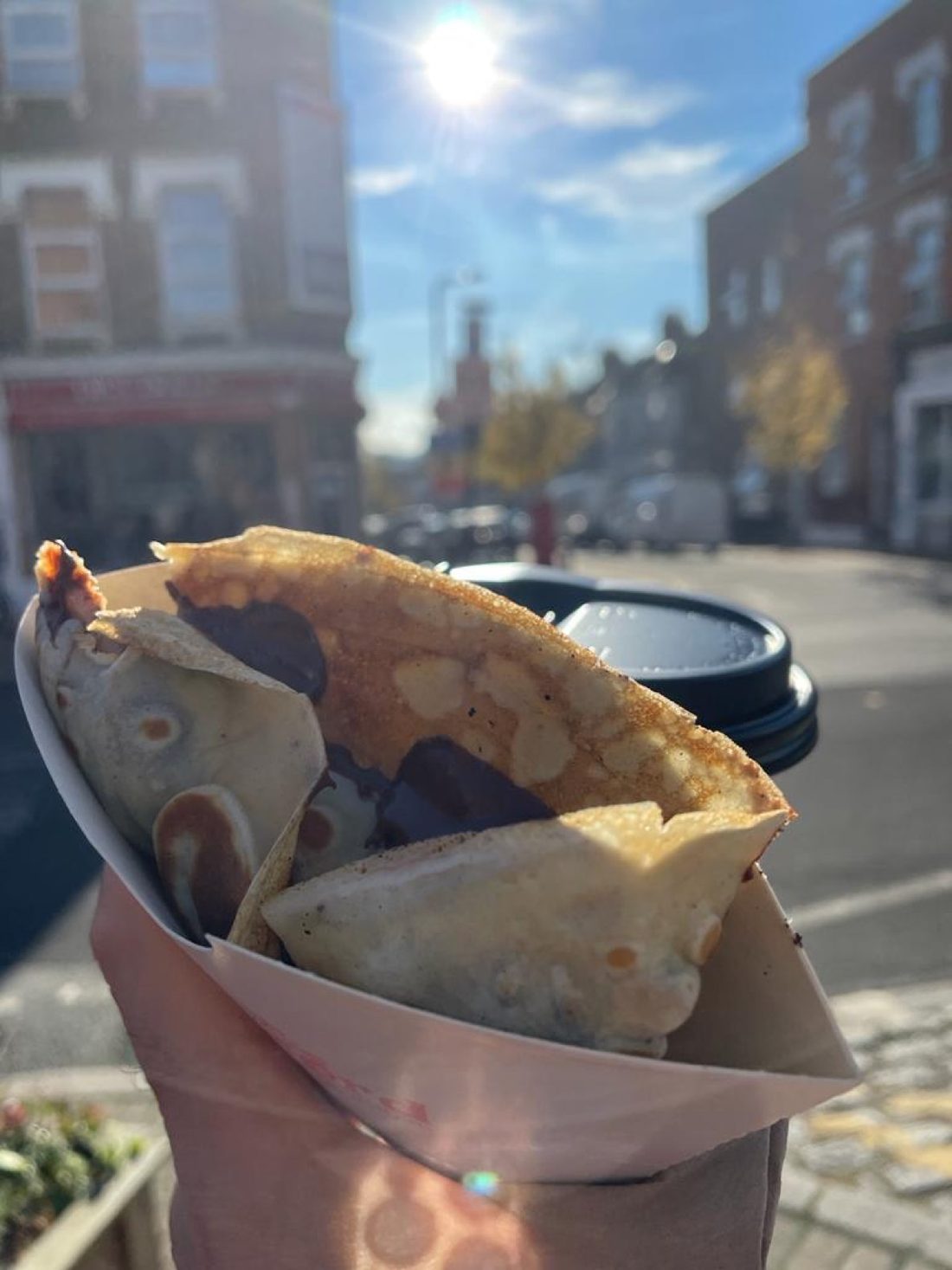 A folded crepe in a paper cone and a takeaway coffee held up on Willesden Lane